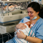 Mother sitting in a hospital chair holding her premature newborn baby wrapped in a blanket in the NICU
