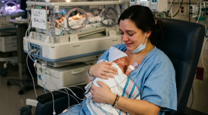 Mother sitting in a hospital chair holding her premature newborn baby wrapped in a blanket in the NICU